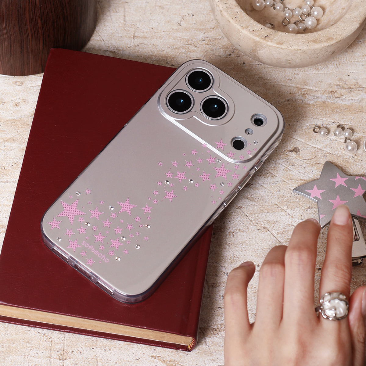 Phone case with pink star designs on a wooden surface with a hand and jewelry.