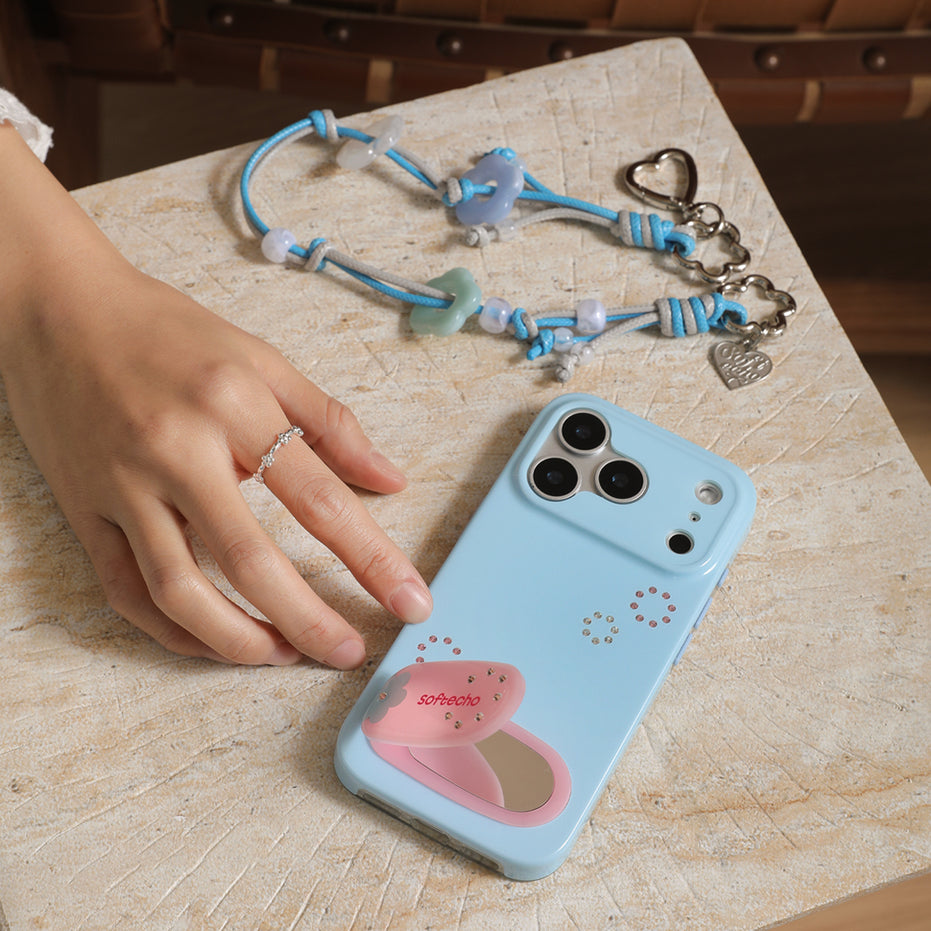 Blue phone case with a pink heart design on a wooden surface, next to a hand and a necklace.