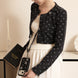 Woman wearing a black patterned jacket holding a phone case with a book in a room.