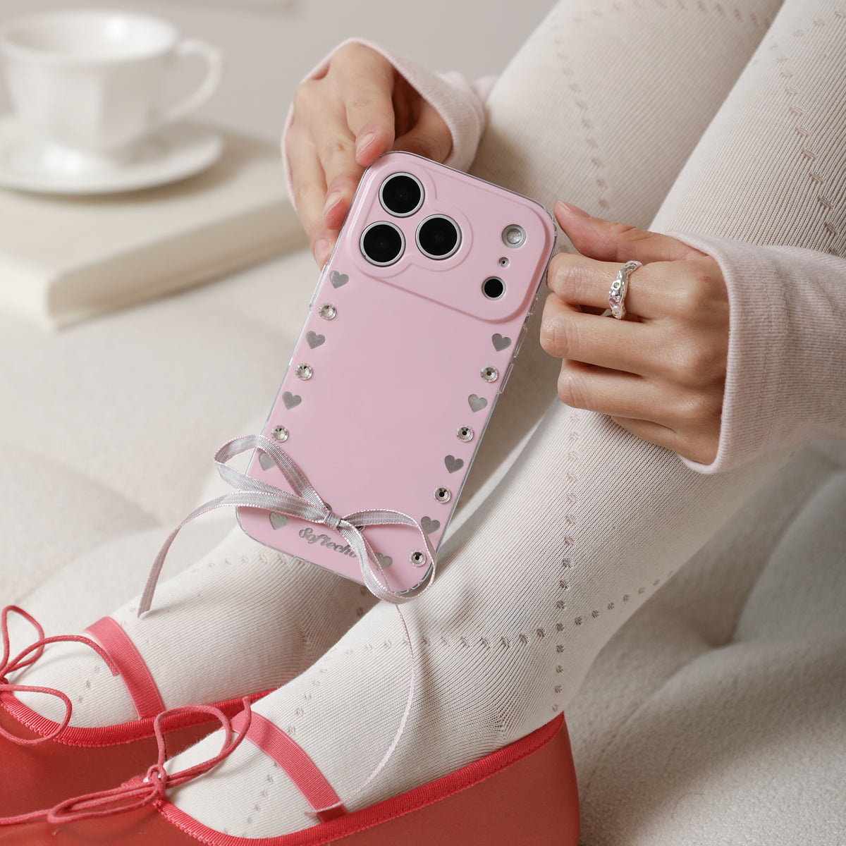 Person holding a pink phone case with heart patterns and a bow, wearing red shoes and a white sweater.