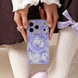 Person holding a phone with a purple floral case, wearing a white lace top and light-colored pants.