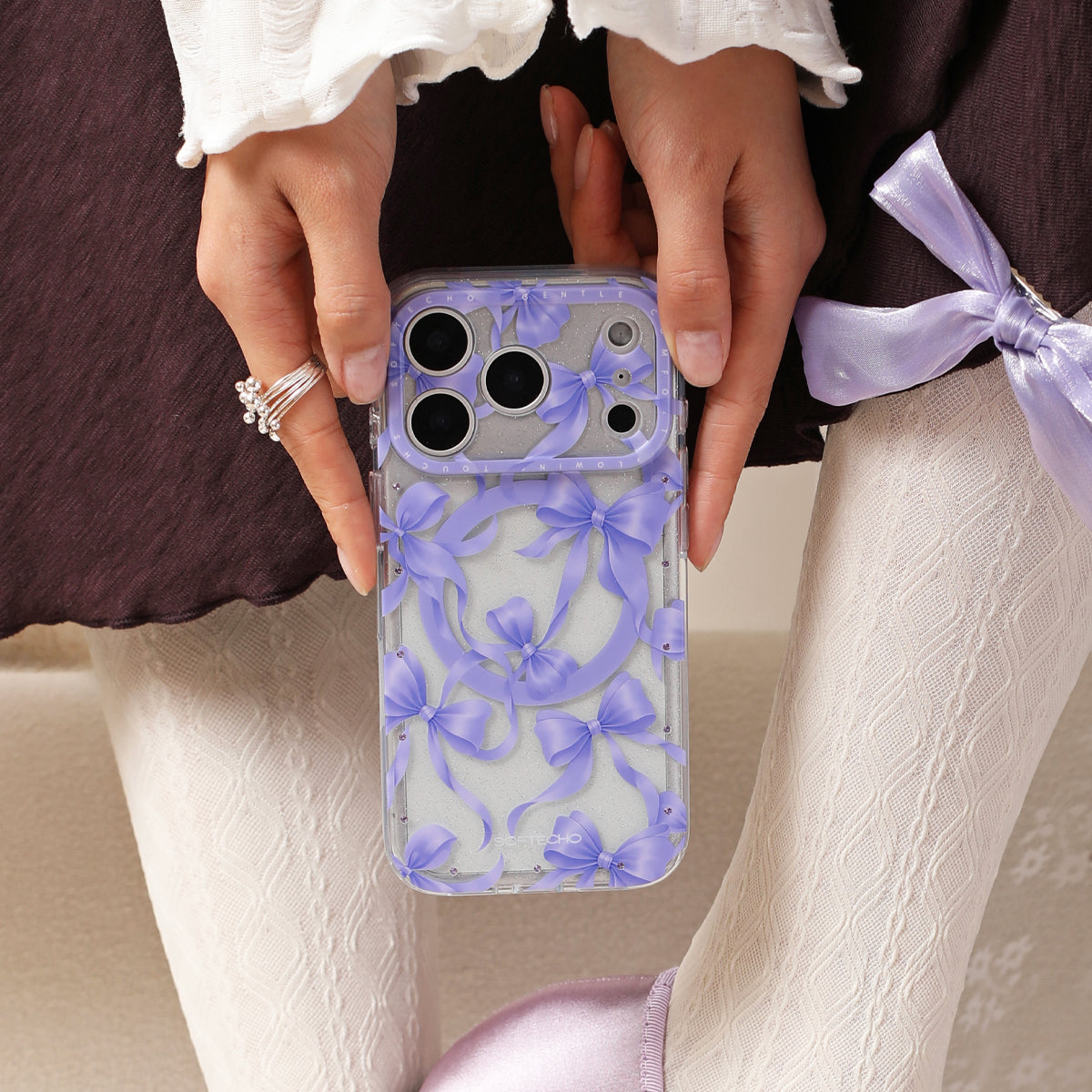 Person holding a phone with a purple floral case, wearing a white lace top and light-colored pants.