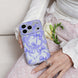 Phone case with purple floral design held by a hand, with a white rose on a light pink fabric background.
